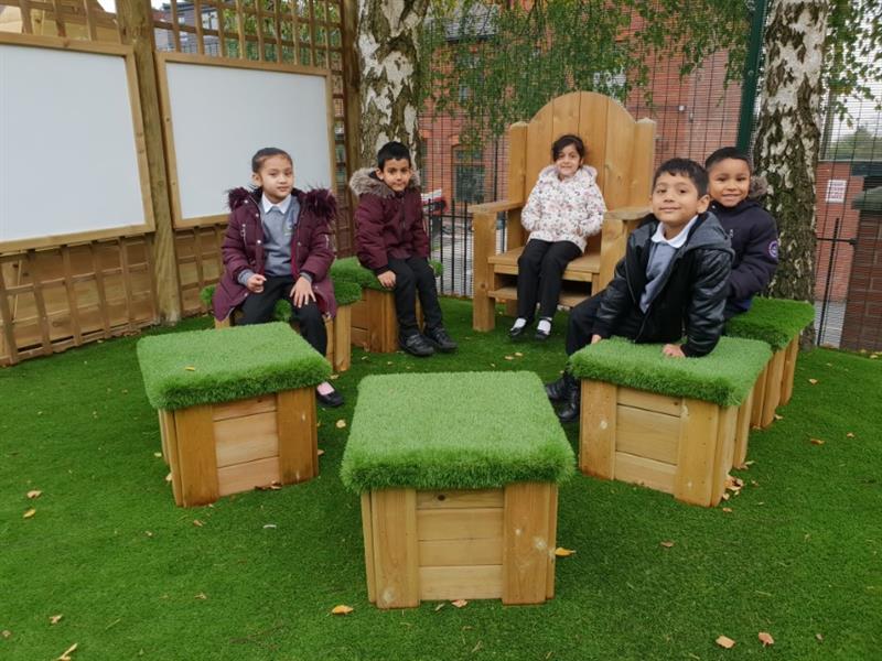 4 children are sat on artificial grass topped seats whilst one child sits on a storytelling chair that have been installed onto artificial grass in front of 2 outdoor whiteboards. 