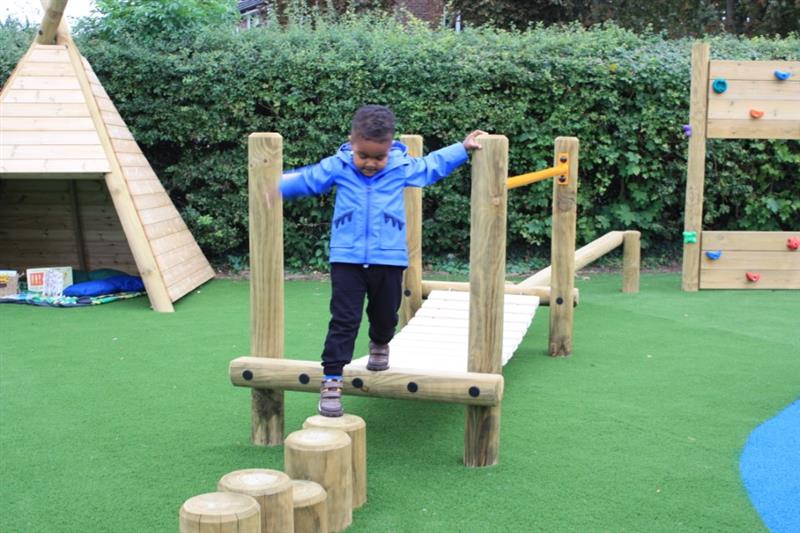 1 boy wearing a blue coat and black trousers stepping off a wobbly bridge onto stepping stones which have been installed onto artificial grass with a wig wam and climbing wall installed behind the bridge. 