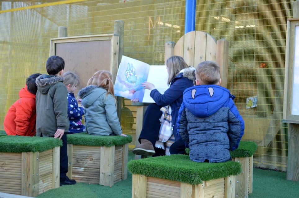 A group of 5 children are sat on top of artificial grass topped seats whilst a teacher sits on a Storytelling chair, carrying out a guided reading lesson in a creative area
