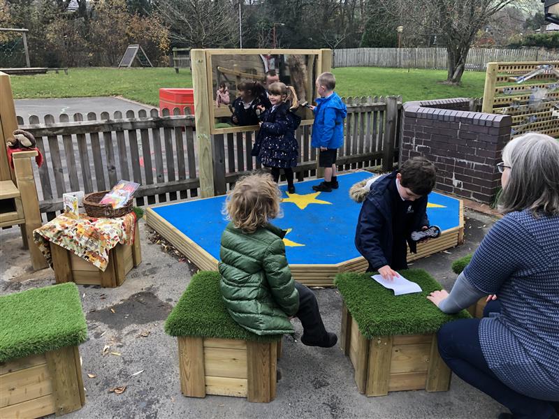 2 children stood on a performance stage topped with blue safeturf and yellow stars in front of playground mirrors whilst 2 children and one teacher sit on artificial grass topped seats in the audience. 