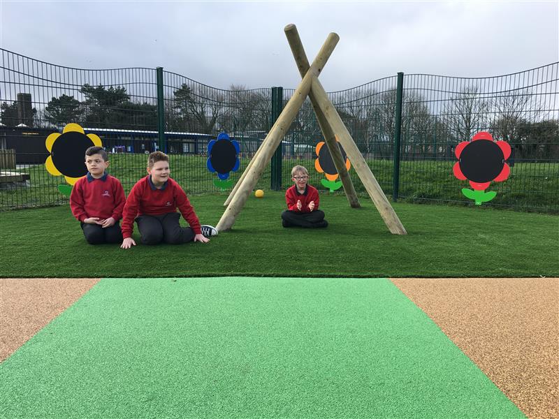 2 children sat next to wigwam posts whilst one child sits underneath the wigwam posts which have been installed onto artificial grass in front of a green fence with 4 different coloured flowers on. 