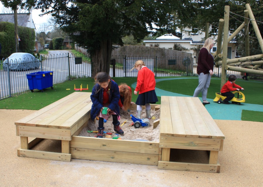 Three children are playing in a sandbox, whilst a teacher stands close by. The teacher is currently watching a child on a tricycle, who is cycling around a wetpour track.