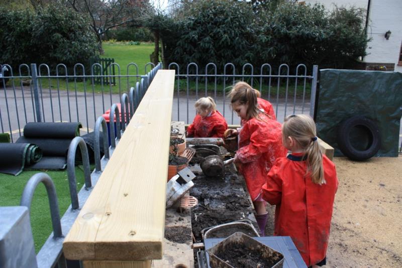children stand around a mud kitchen and play with mud whilst wearing red aprons to protect their clothes