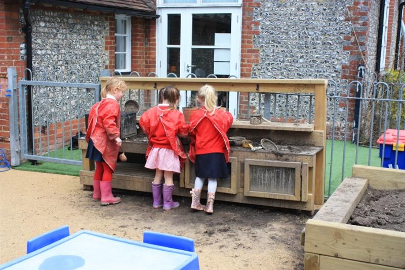 3 children gathered around a mud kitchen using metal bowls to mix their ingredients and putting cake tins in the oven of the kitchen
