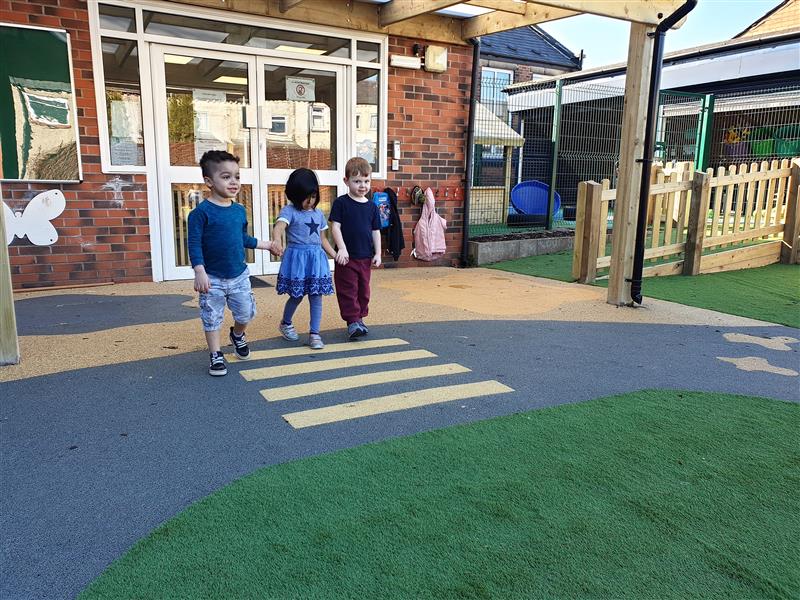 3 children, 2 boys and 1 girl holding hands as they cross a zebra crossing which has been created using playground markings and placed in front of the school building. 