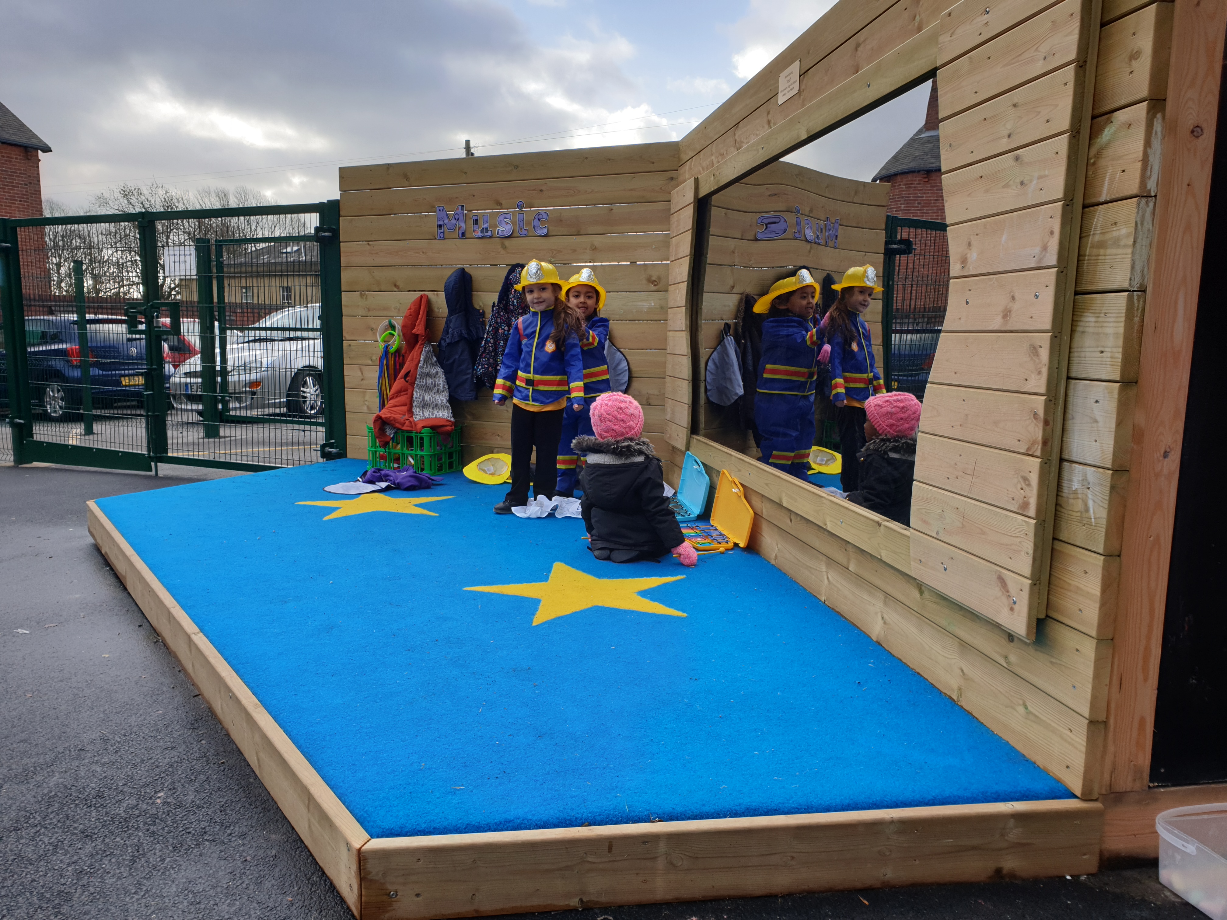 2 children are dressed up as fireman, wearing blue jackets and yellow fireman hats. One child is sat next to them and is looking at some instruments. The back of the stage is wooden, with one part having a mirror attached to it. The surface of the stage is blue saferturf, with yellow stars.