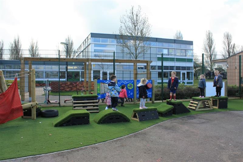 5 children playing on Get Set Go! blocks which have been installed onto artificial grass near to the school building, with two children running past behind the blocks. 