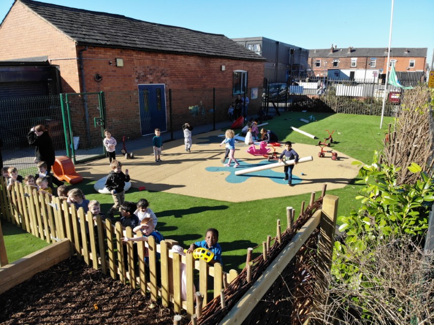 An artificial grass and wetpour playground with freestanding playground equipment placed on it, with a large group of children playing there.