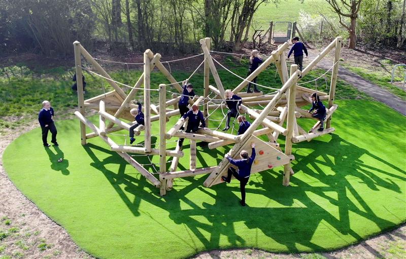 10 children playing on a large climbing frame with a rock climbing wall which has been installed onto artificial grass in a forest area in front of large trees with one child sat on the grass watching the other children play. 