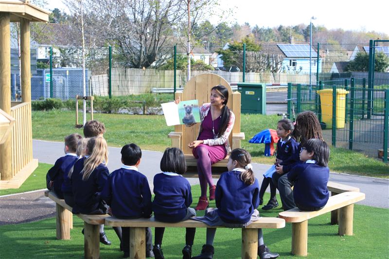 children sit gathered in a semi circle on perch benches facing a storytelling chair whilst a teacher in a floral cardigan sits on the storytelling chair and reads them a story