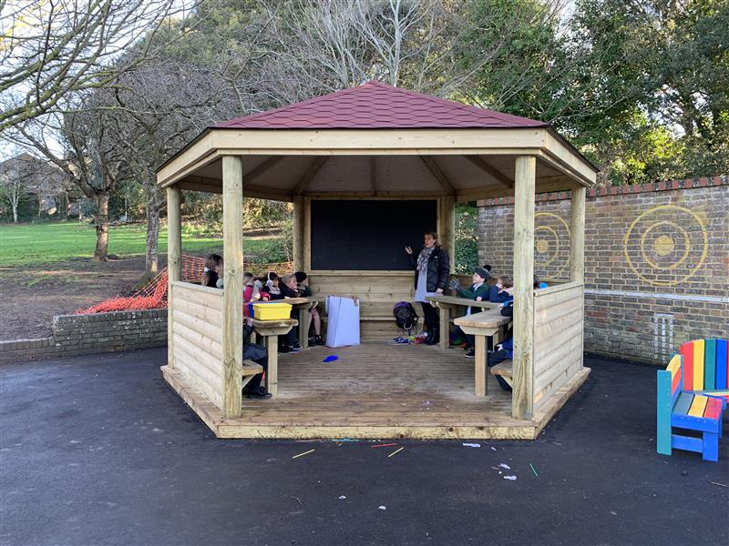 A class of children sat inside of a hexagonal gazebo with a large chalkboard whilst one teacher stands next to the chalkboard teaching a lesson. The gazebo has been installed next to a multi-coloured bench. 