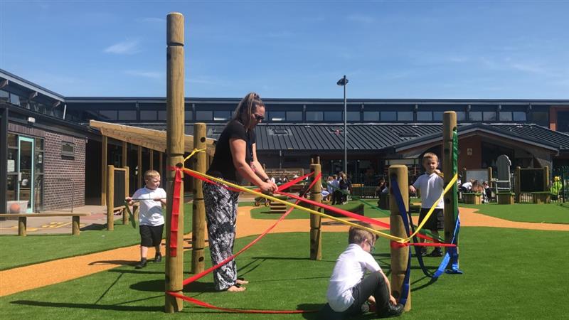 3 children and 1 teacher using red, yellow, green and blue ribbon to wrap around 5 timber poles that have been installed onto artificial grass in the middle of the playground. 