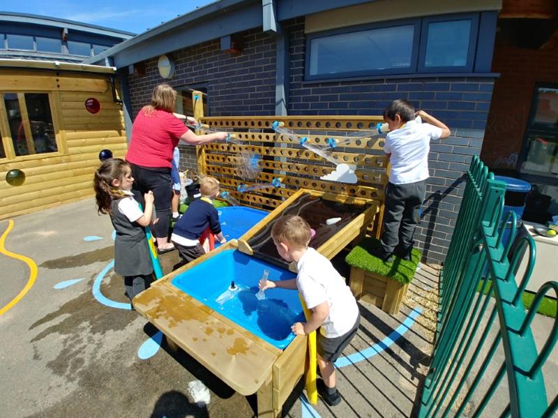 Four children playing on the water wall and damming station, two teachers are giving them support