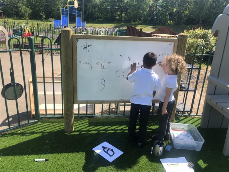 2 children wearing white tops and grey trousers are stood at a whiteboard outside drawing pictures. The whiteboard has been installed onto artificial grass.