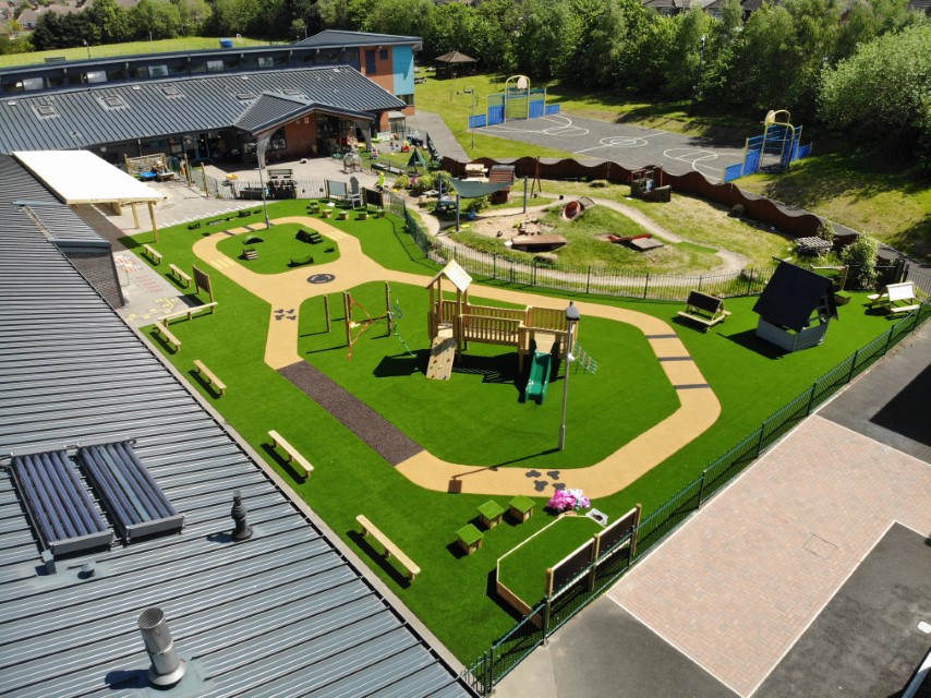 A play area that has an artificial grass surface installed. A playtower, den posts and get set go blocks can be seen across the area. A wetpour path has been installed on the surface.