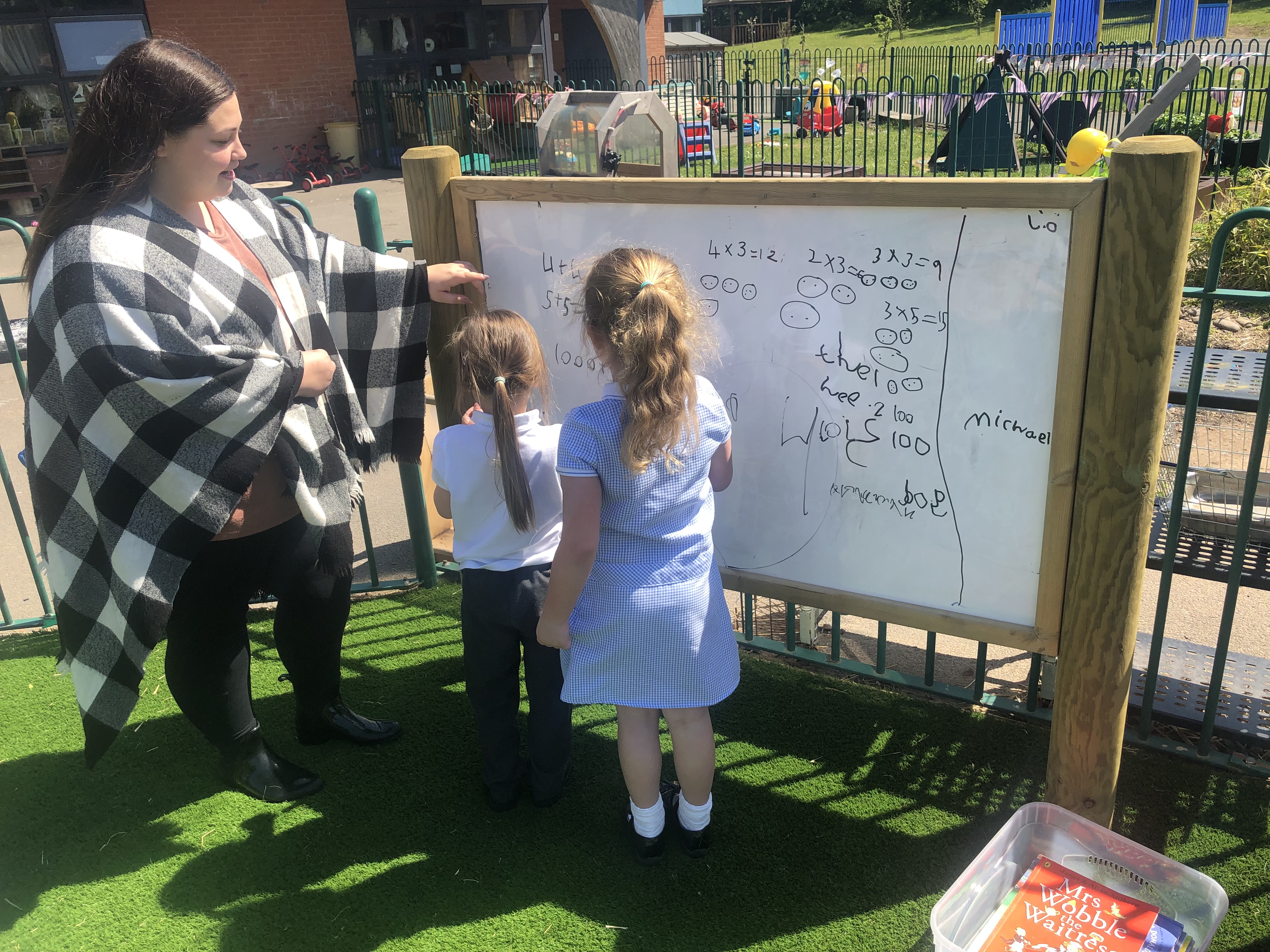 A teacher is guiding two students who are writing on a whiteboard. The two little girls are working out maths problems