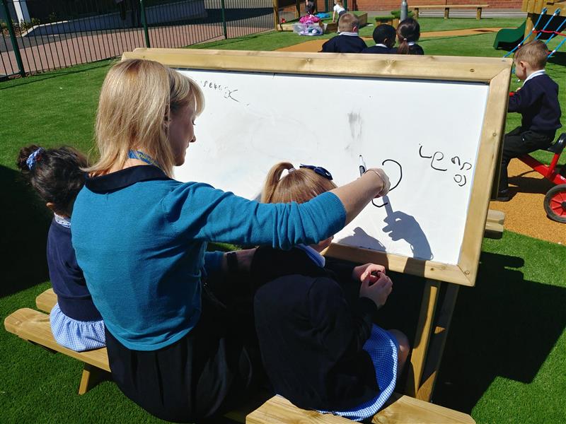 children sit either side of the teacher on the easel table and draw on the whiteboard with black pens