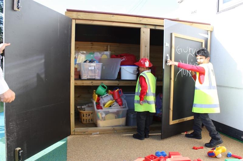 Child opening the doors of a large playground storage unit whilst another child looks inside