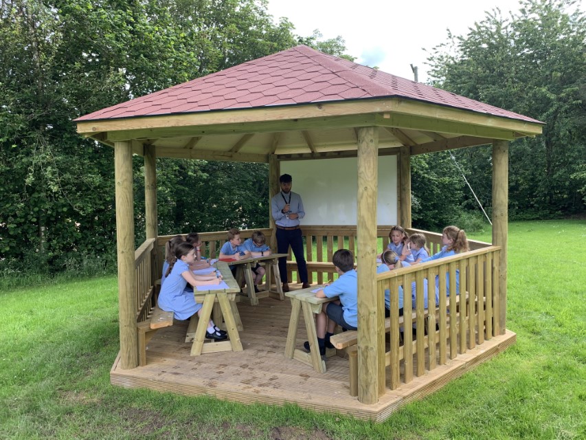 A teacher is stood next to a whiteboard that has been installed inside a wooden gazebo, with a class full of children all sat on benches. Tables are set infront of the children and they are currently working on a project as the teacher is teaching a subject.