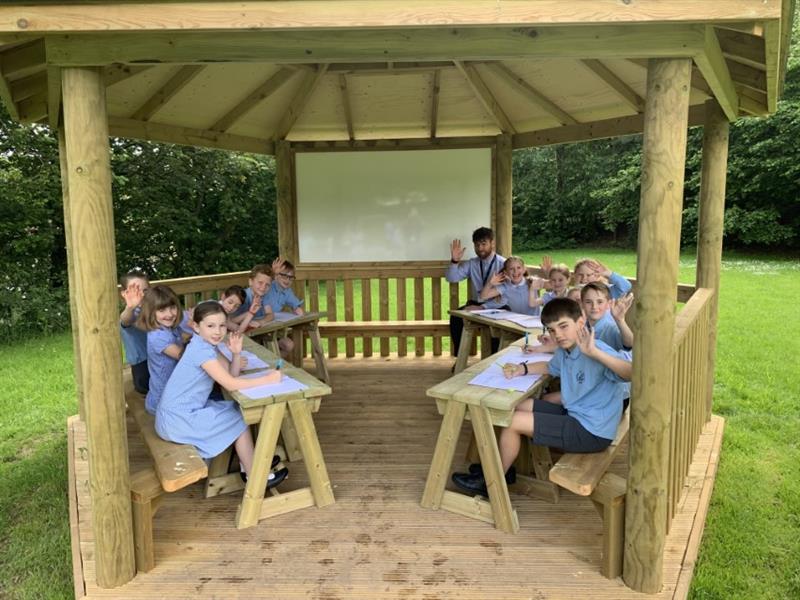 children in blue school uniform sit inside th gazebo classoom and face the camera smiling at it as they work on desks in front of them