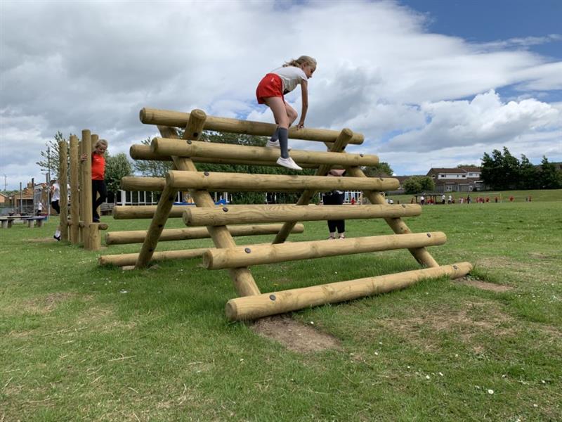 One girl wearing red shorts and a white top is climbing over a timber frame as part of an obstacle course which has been installed onto the school field.