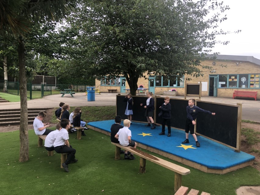 4 children are stood on an outdoor stage, performing a dance to an audience of other children. The audience are sat on long wooden benches, looking at the stage. The surface of the stage is a light blue saferturf, with yellow stars showing on it. Chalkboards can be seen behind the performers.