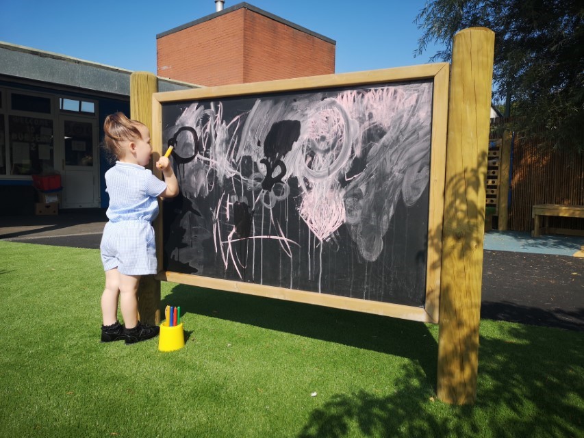 A little girl holding a paint brush and is painting water onto a huge outdoor chalkboard covered with pink chalk. She is painting shapes and letters.