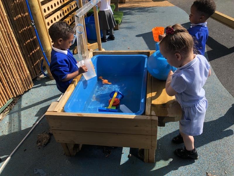 3 children playing in the sunshine with a blue water table using jugs and sand castle buckets to play with the water.