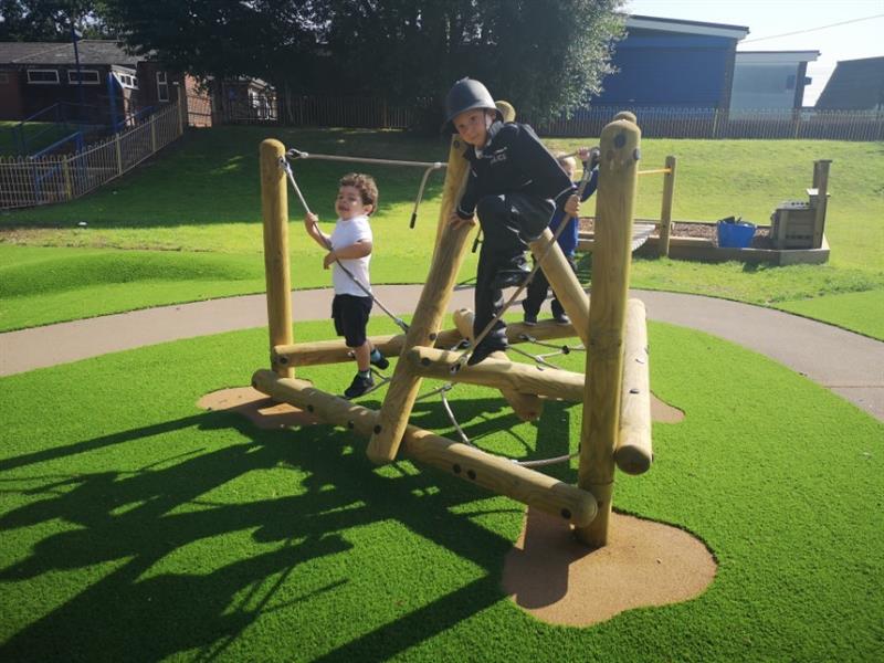 2 children on a climbing frame, one dressed as a policeman wearing a police hat and the other child is wearing a white t-shirt and shorts. The climbing frame has been installed onto artificial grass.