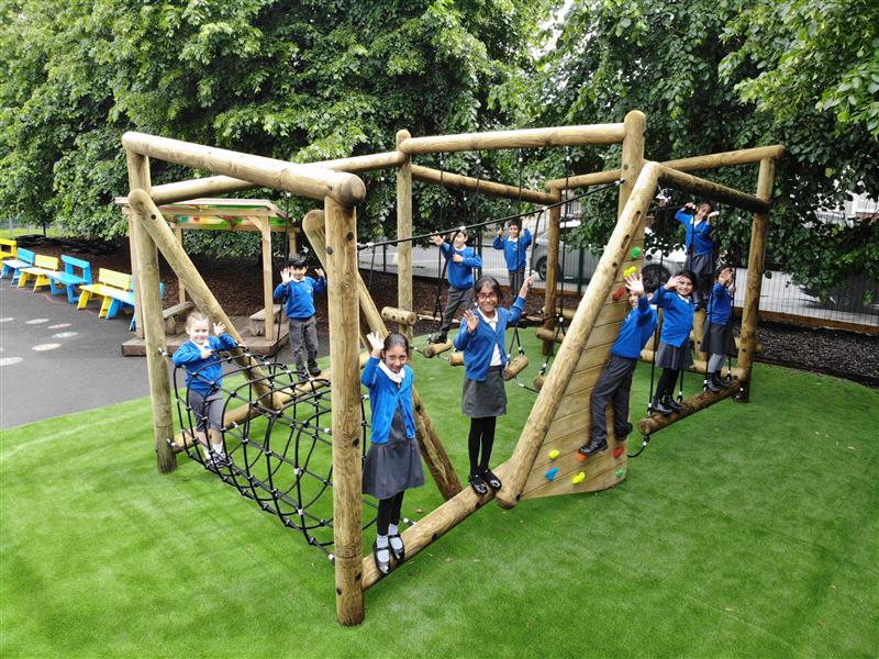 9 children playing on a large climbing frame which has been installed onto artificial grass, all the children are stood waving towards the camera, wearing blue school jumpers. 