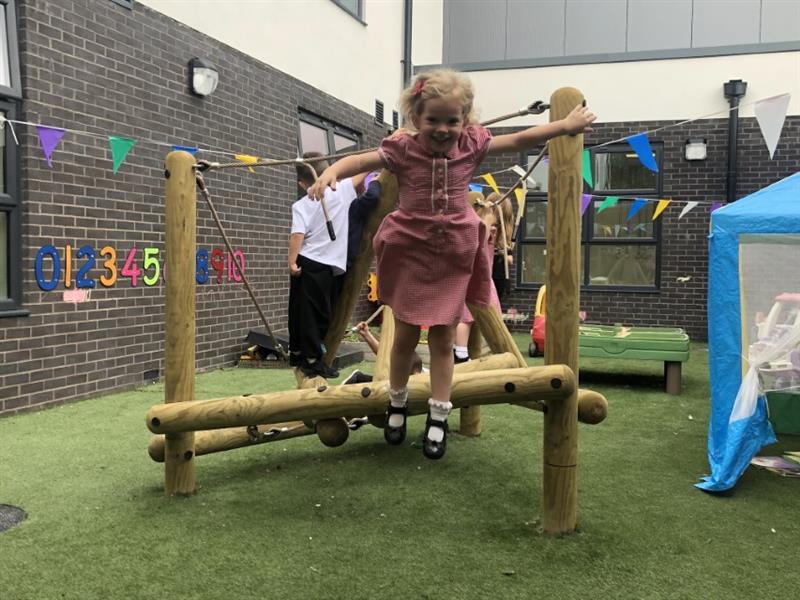 A girl with blonde hair wearing a red summer school dress jumping off a climbing frame that has been installed onto artificial grass placed in front of the school building. 