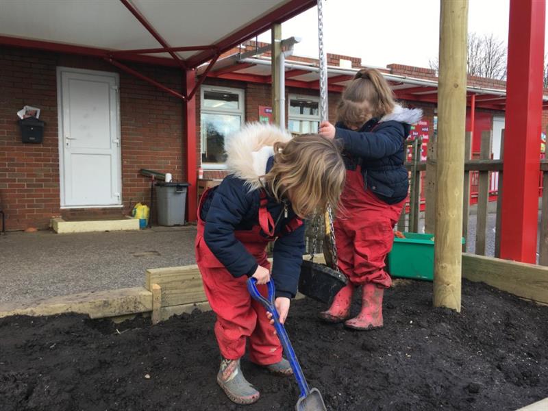 2 girls wearing red overalls and wellington boots stood inside of a mud pit playing with a blue spade and a rope and pulley feature to move the mood around. 