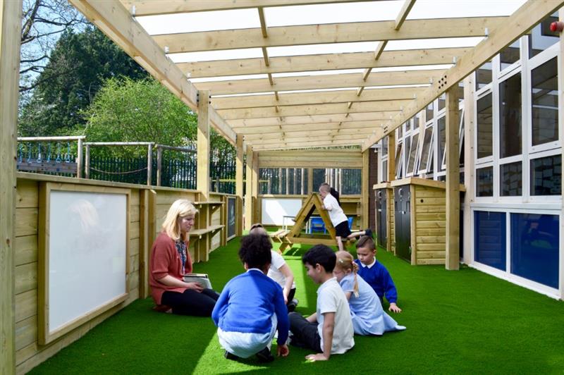 children sit in a semi circle on the artificial grass surfacing within the timber canopy in front of a teacher in a pink cardigan