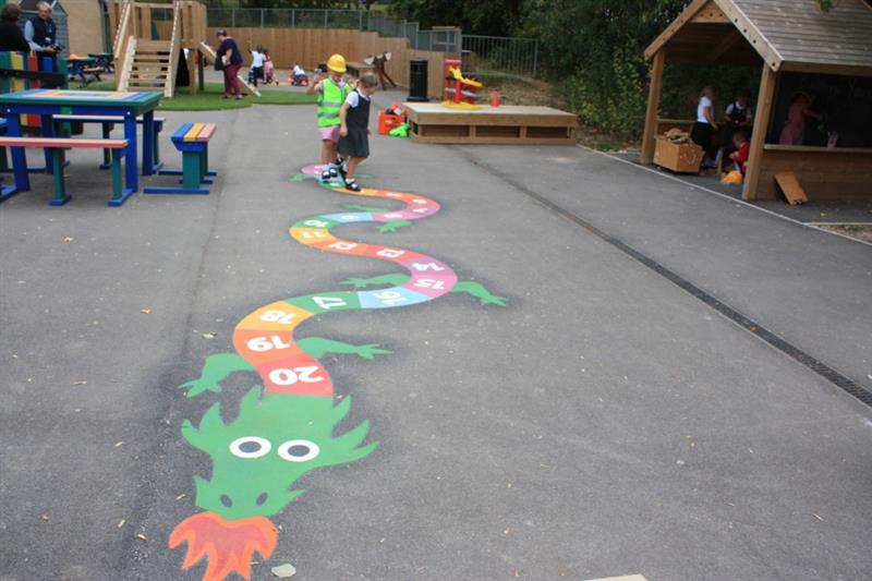 2 children playing on a mmulti-coloured snake with numbers playground marking, one girl wearing a red dress, bright jacket and yellow builders hat whilst the other girl wears a grey dress. 