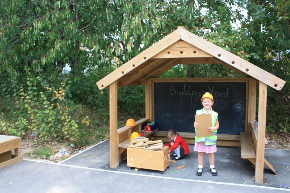 2 children are stood inside the Giant Playhouse as they take part in pretend play. One child has dressed up as a builder as the other child looks through boxes for building materials.