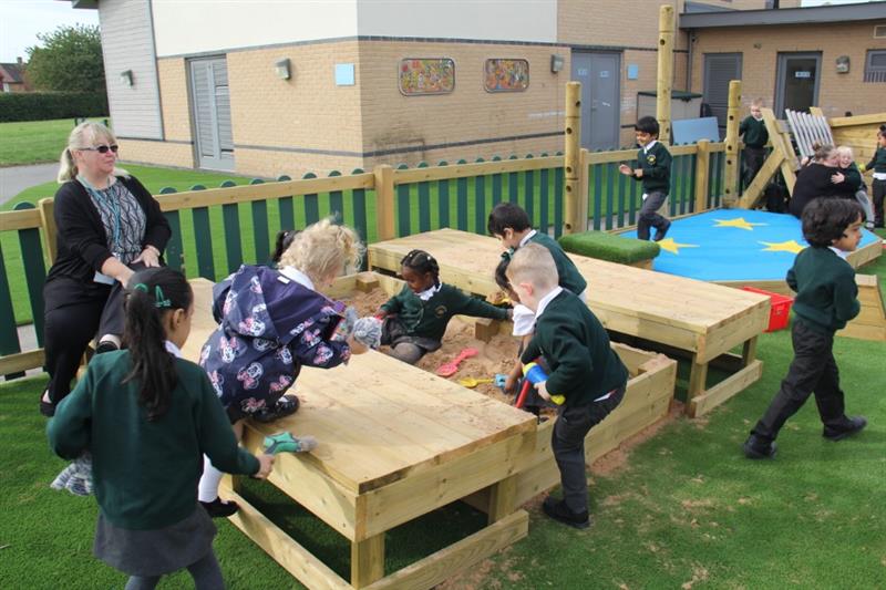 children in green school uniform sit in the large sand box and play with the sand and the tools to move the sand such as diggers and spades