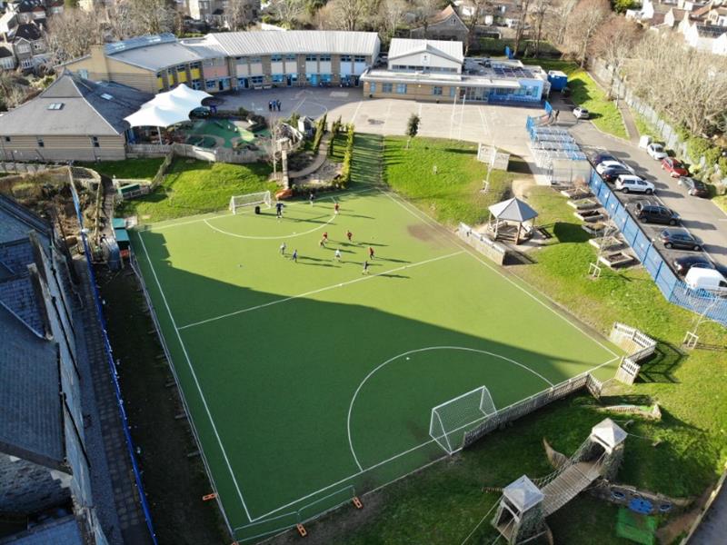 Aerial view of huge MUGA pitch witch children playing on one half of the pitch in the sunshine. The MUGA has been installed next to the school building. 