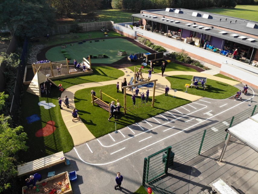 A playground that has a variety of surfacing types present on it. There's a wetpour section with get set go blocks on it, an artificial grass surface with wooden play equipment installed on it and a tarmac playground with road line markings on it.