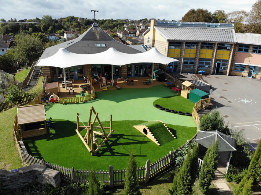 A play area that is shown from the air. The play area is has artificial grass surfacing, with green wetpour surfacing. A Play Frame, a play house and an Up and Under Block can be seen, with other pieces of play equipment and outdoor storage options being seen.