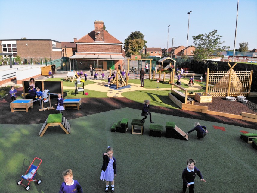 A drone shot showcasing a school playground and all of the children playing on it. Different pieces of play equipment can be seen throughout the area, with children safely taking part in play.