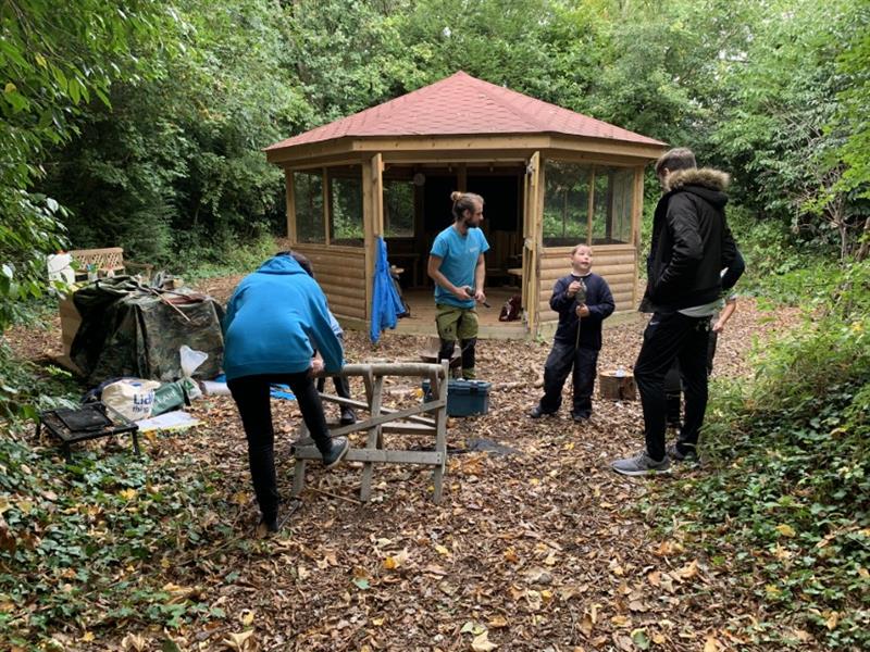 3 children talking to 3 installers in a school forest surrounded by large trees with a large bespoke hexagonal gazebo in the middle of the forest. 