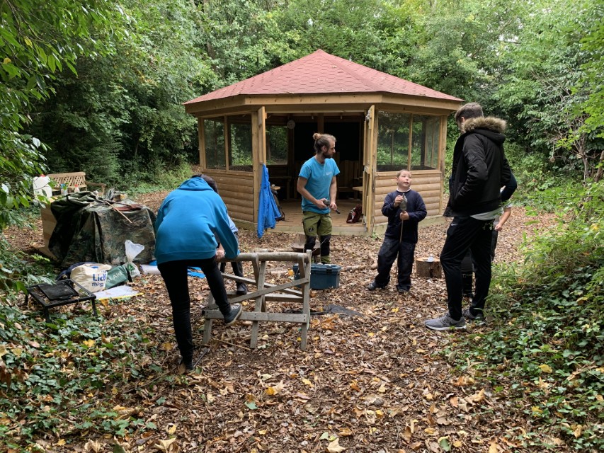 A group of people are stood out the front of a 5M Hexagonal Gazebo, made by Pentagon Play. The gazebo has been installed in a forest and is surrounded by trees and foilage.