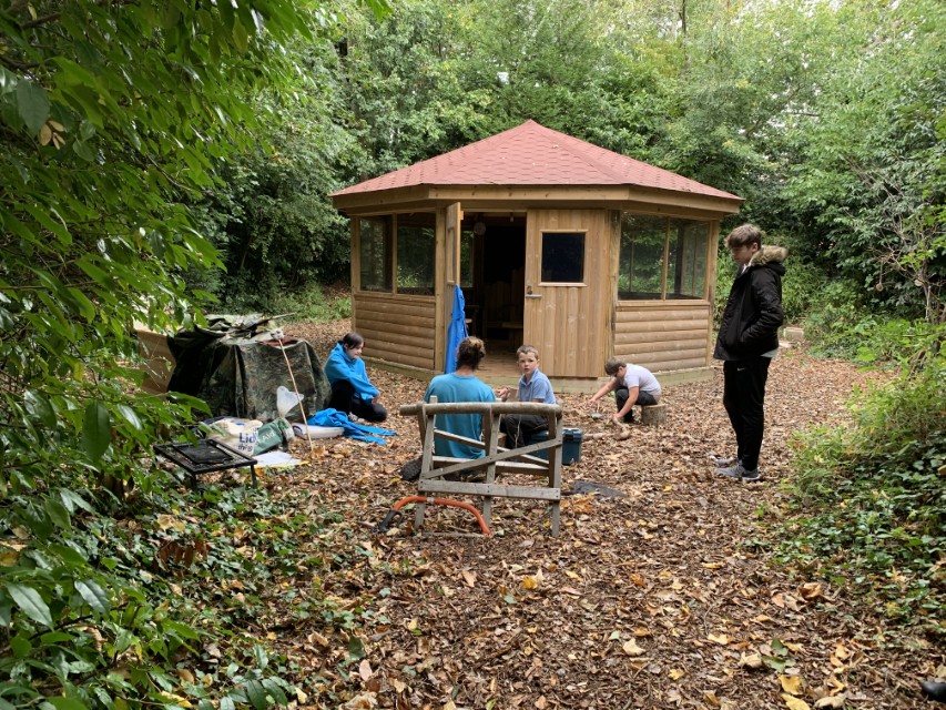 A gazebo that has been installed in the middle of a forest, with dried leaves covering the floor. The children are playing with the leaves and creating things with the materials that surround them.