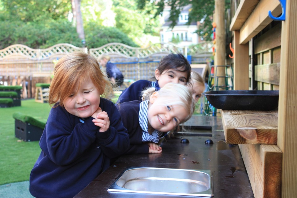 Three children are leaning against a Mud Kitchen and are smiling directly into the camera.