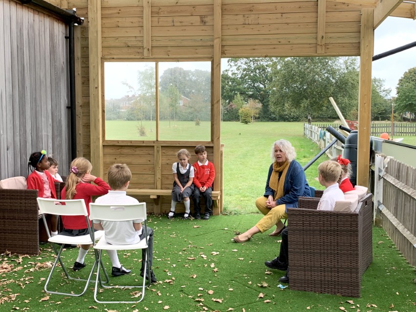 A teacher is sat on a chair and is talking to her class. A group of children are all sat on different types of seating options as they look at the teacher. The class is taking place outside and underneath a canopy.