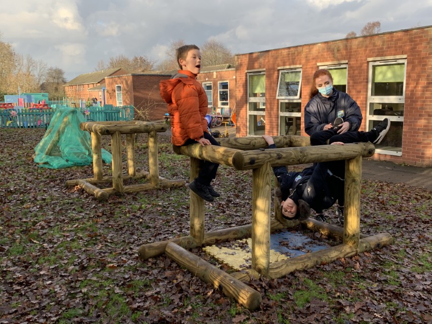 A SEN child is sat on a wooden structure as he smiles, whilst another boy is hanging from the structure by his legs. A teaching assistant is standing nearby to protect the boys.