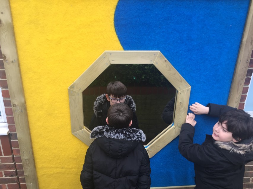 Two little boys are playing with a Sensory Panel, with one boy looking into the mirror and another boy stroking the blue grass beside it.