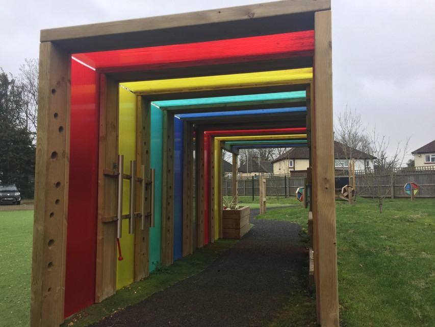 A square-shaped tunnel that has a variety of coloured plastic panels around it. Inside the tunnel is a variety of sensory play equipment with a planter on the other side of the tunnel. The tunnel has been installed on natural turf with a wetpour path going through it.