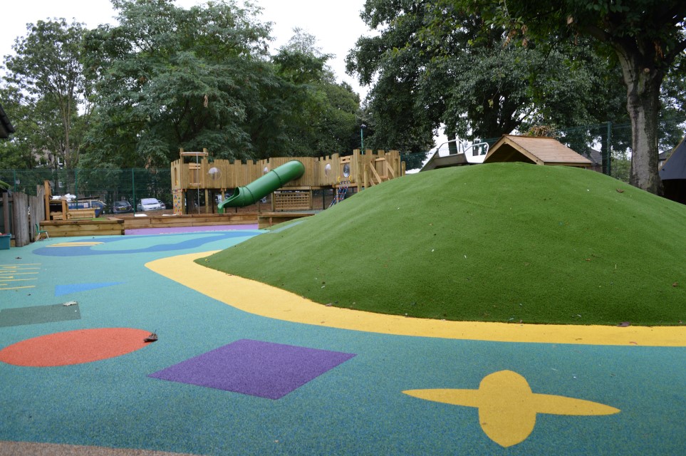 A wetpour playground with a blue base. There are shapes of flowers, squares, circles, triangles and numbers. A giant artificial grass hill is in the middle of the playground, with a play tower in the background