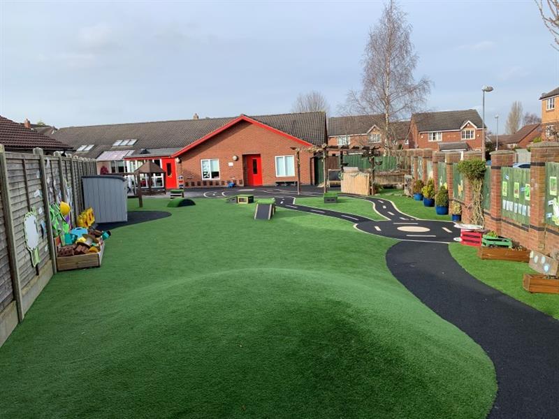 School playground with a large mound covered with artificial grass next to a path leading to a roadway and get set go! blocks at the other side of the playground, in front of the school building which has red doors. 